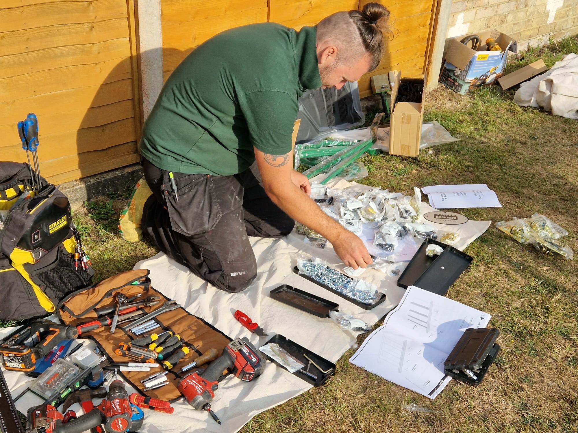 A tradesman kneeling on a dust sheet in a garden, sorting the parts of a swing set kit with his tools laid out